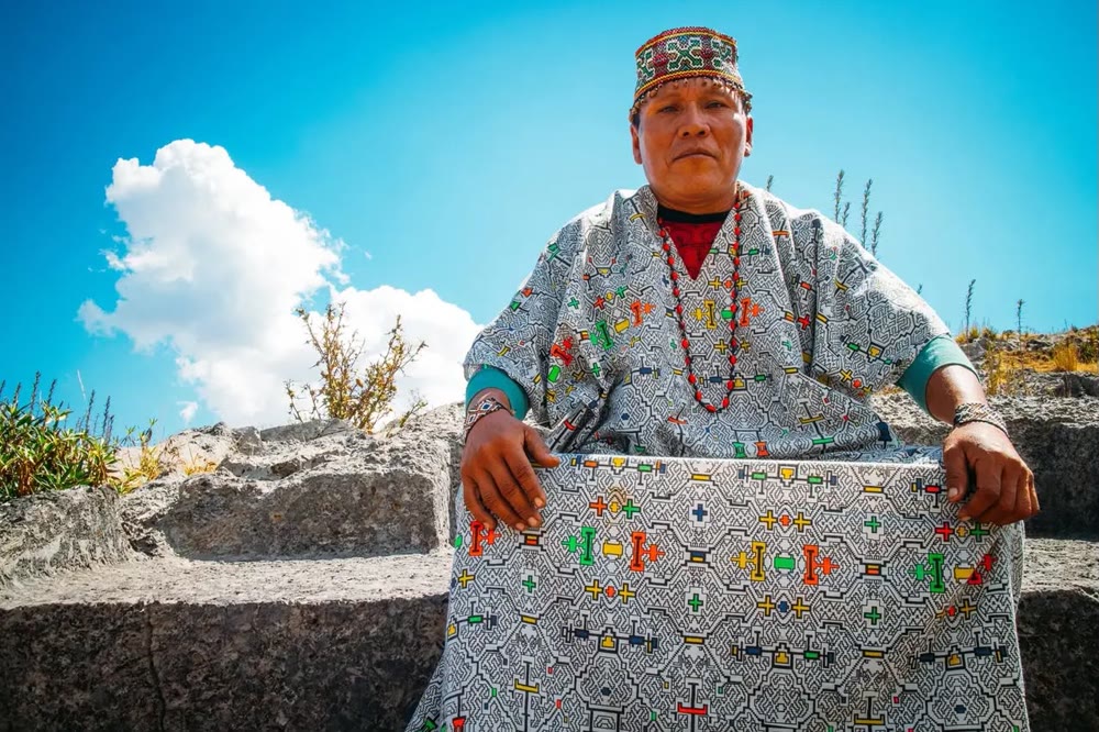 A Shipibo maestra in traditional embroidered dress and a beaded corona, seated outdoors in Peru