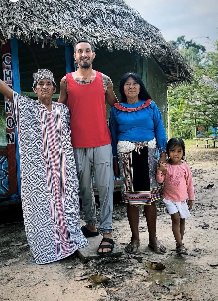 A Shipibo elder in traditional dress beside a facilitator outside a thatched maloca in Peru